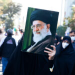 Regime Change FAILING—CIA Veterans Issue Dire Warning… Person holding a large portrait in a crowd