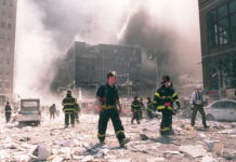 Firefighters walking through debris near a heavily damaged smoking building