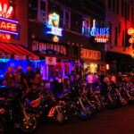 RECALL: Manufacturing Flaw Turns Harleys Into Time Bombs… Vibrant street scene at night with neon signs and parked motorcycles