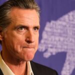 A serious-looking political figure at a press event with a bear graphic in the background