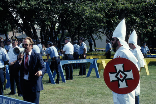 shutterstock_711532204.jpg Police monitor a man speaking and a person in KKK outfit.