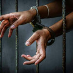 Handcuffed hands reaching through jail cell bars.