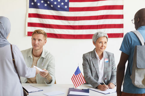 shutterstock_1810359100 Election workers assist voters at a registration table with a U.S. flag behind them