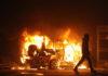 Burning vehicle at night, silhouetted person walking past.